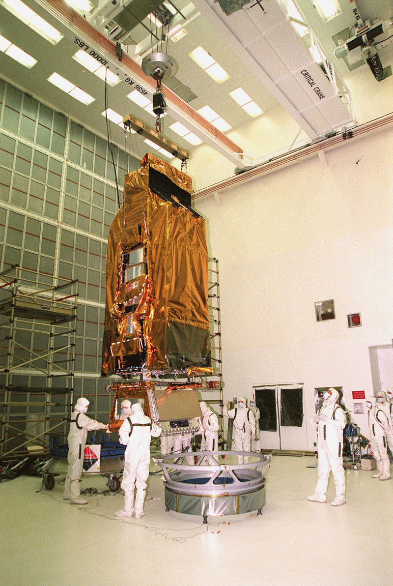 While a crane lifts NASA’s FUSE satellite, workers at Hangar AE, Cape Canaveral Air Station, help guide it toward the circular payload-attach fitting in front of it.