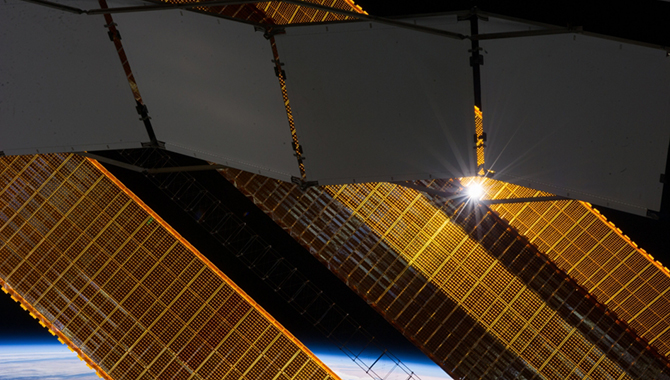 A tiny representation of the sun sneaks through between a truss-based radiator panel and a primary solar array panel on the Earth-orbiting International Space Station in this photograph taken by one of the Expedition 38 crew members on Jan. 2, 2014. Clouds over Earth and the blackness of space share the background scene.