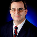 Portrait from the shoulders up of JD Polk. He is wearing glasses, a dark suit, and a striped burgundy tie.