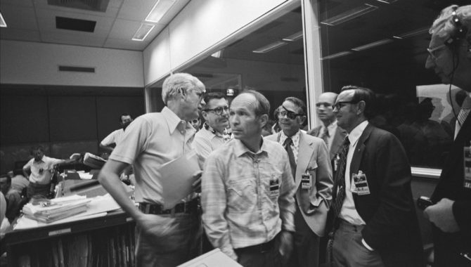 At the Johnson Space Center (JSC) Mission Operations Control Center, NASA officials discuss the problems with the micrometeroid shield on Skylab. From left to right, they include Jack Kinzler, whose Skylab sunshield solution earned him the NASA Distinguished Service Medal, along with William Schneider, Maxim Faget, Dale Myers, JSC Director Chris Kraft, and Kenneth Kleinknecht. Credit: NASA