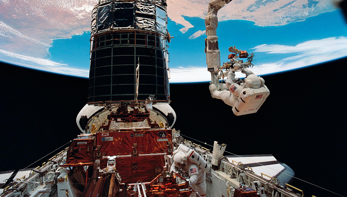 Astronaut F. Story Musgrave, anchored on the end of the Remote Manipulator System (RMS) arm, prepares to be elevated to the top of the towering Hubble Space Telescope (HST) to install protective covers on magnetometers. Astronaut Jeffrey A. Hoffman (bottom of frame) assisted Musgrave with final servicing tasks on the telescope, wrapping up five days of extravehicular activities (EVA). Credit: NASA