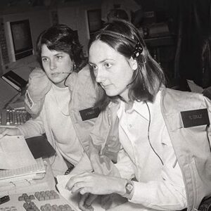 A historical black and white photo of two young women, Michele Brekke and Susan Creasy, who were working in the shuttle mission simulator. To keep warm, they were wearing coats from the STS-2 crew–Joe Engle and Dick Truly. This image is used as a cover for the book “Making Space for Women” by Jennifer Ross-Nazzal. The image is symbolizing that women became accepted and a part of NASA’s culture as the workforce became more diverse. Credit: NASA