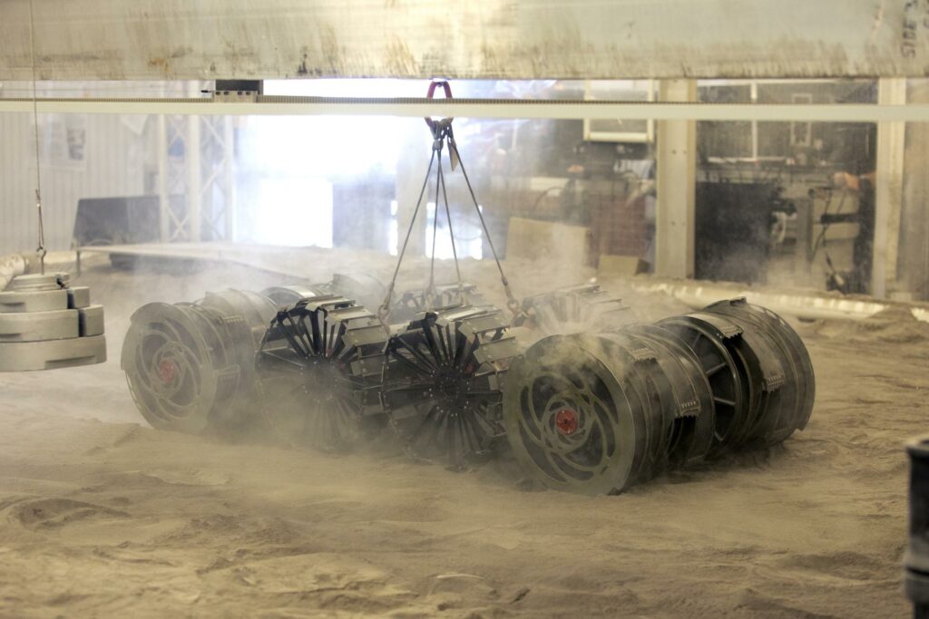 A team from the Granular Mechanics and Regolith Operations Lab tests the Regolith Advanced Surface Systems Operations Robot (RASSOR) in the regolith bin inside Swamp Works at NASA’s Kennedy Space Center in Florida on June 5, 2019. Tests use a gravity assist offload system to simulate reduced gravity conditions found on the Moon. On the surface of the Moon, mining robots like RASSOR will excavate the regolith and take the material to a processing plant where usable elements such as hydrogen, oxygen and water can be extracted for life support systems. RASSOR can scoop up icy regolith which can be used to make operations on the Moon sustainable. Credit: NASA/Kim Shiflett