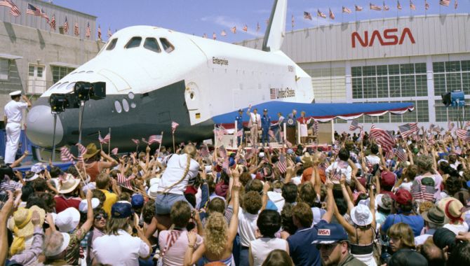 President Ronald Reagan (tan suit) waves from the podium as thousands of spectators cheer. More than half a million people gathered to watch Space Shuttle Columbia touch down following STS-4. The prototype Space Shuttle Enterprise is behind Reagan. Photo Credit: NASA