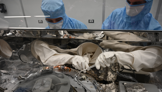 Astromaterials processors Mari Montoya, left, and Curtis Calva, right, use tools to collect asteroid particles from the base of the OSIRIS-REx science canister. NASA has already collected 70.3 grams of material with more still inside the canister. Photo Credit: NASA