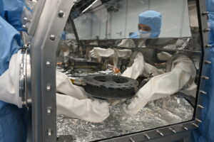 Astromaterials processor Mari Montoya and OSIRIS-REx curation team members set the TAGSAM (Touch and Go Sample Acquisition Mechanism) down in the canister glovebox after removing it from the canister base and flipping it over. Photo Credit: NASA