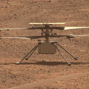 Ingenuity sits on Mars. It resembles a small box with four landing legs and rotors on top. The terrain is brownish-red with scattered pebbles and rocks.