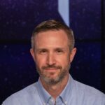 Portrait shot of Jake Bleacher in a blue button-down shirt with the NASA emblem on it. He is sitting at a dais with a transparent surface. A black conference microphone sits in front of him.