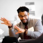 Candid photo of Dr. Ron Gamble. His arms are outstretched to the side as if he is describing something. He has dark hair and wears glasses and a gray shirt with a tie.