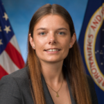 Dr. Mary Skow, female, sitting staring at the camera with a NASA flag and a US flag behind her.