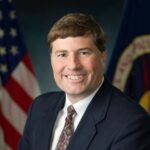 A man smiles for a portrait. He wears a dark blue suit with a white shirt and brown-patterned tie. Two flags are draped behind him, one being the American flag and the other being the NASA flag, which has a blue field with a circular NASA emblem. Credit: NASA