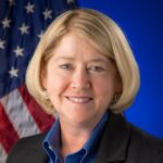 Portrait of Pam Melroy. She smiles and has short, blonde hair. An American flag is draped behind her.