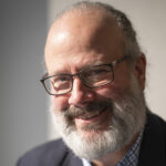 Portrait of Bill Ingalls from the shoulders up. He wears a dark blazer and a button down shirt. He has a beard and wears glasses.