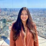 A woman stands on a building's observation deck with glass behind her. A sprawling city is below. She wears a brownish-red jacket and her brown hair goes well past her shoulders. Credit: Sue Kaur