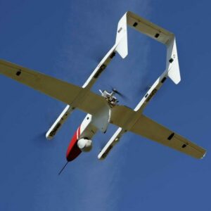 Photo from below of a drone flying against a deep-blue sky. The drone is white with a pointed red nosecone. Credit: Don Richey