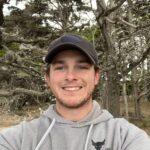 Patrick Hill takes a selfie in a wooded area. He wears a dark gray baseball cap and a gray sweatshirt.