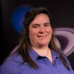 Portrait of a woman smiling in front of a graphic of the planet Saturn. She smiles and is wearing a purple top. Her hair goes a few inches beyond her shoulders.